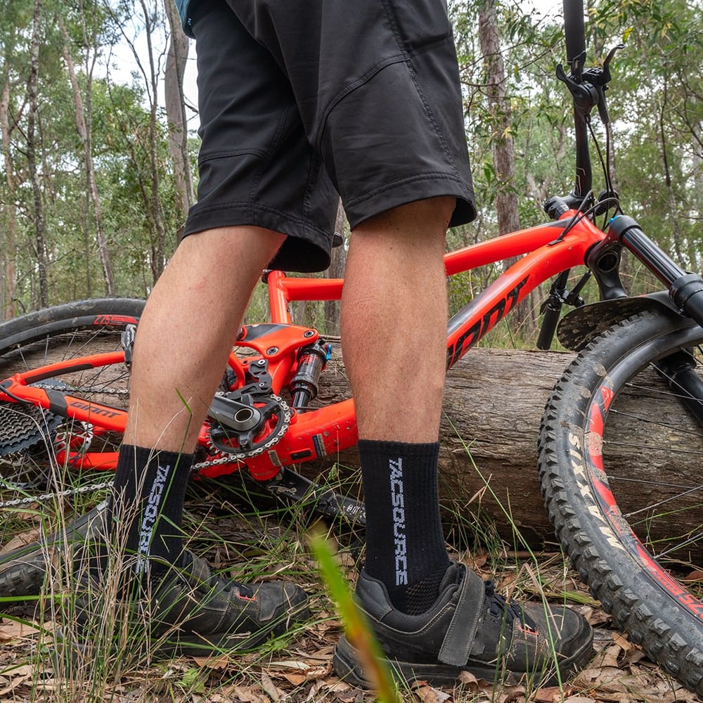 TacSource Active Train 2.0 Socks on a cyclist next to a red mountain bike in a forest setting.