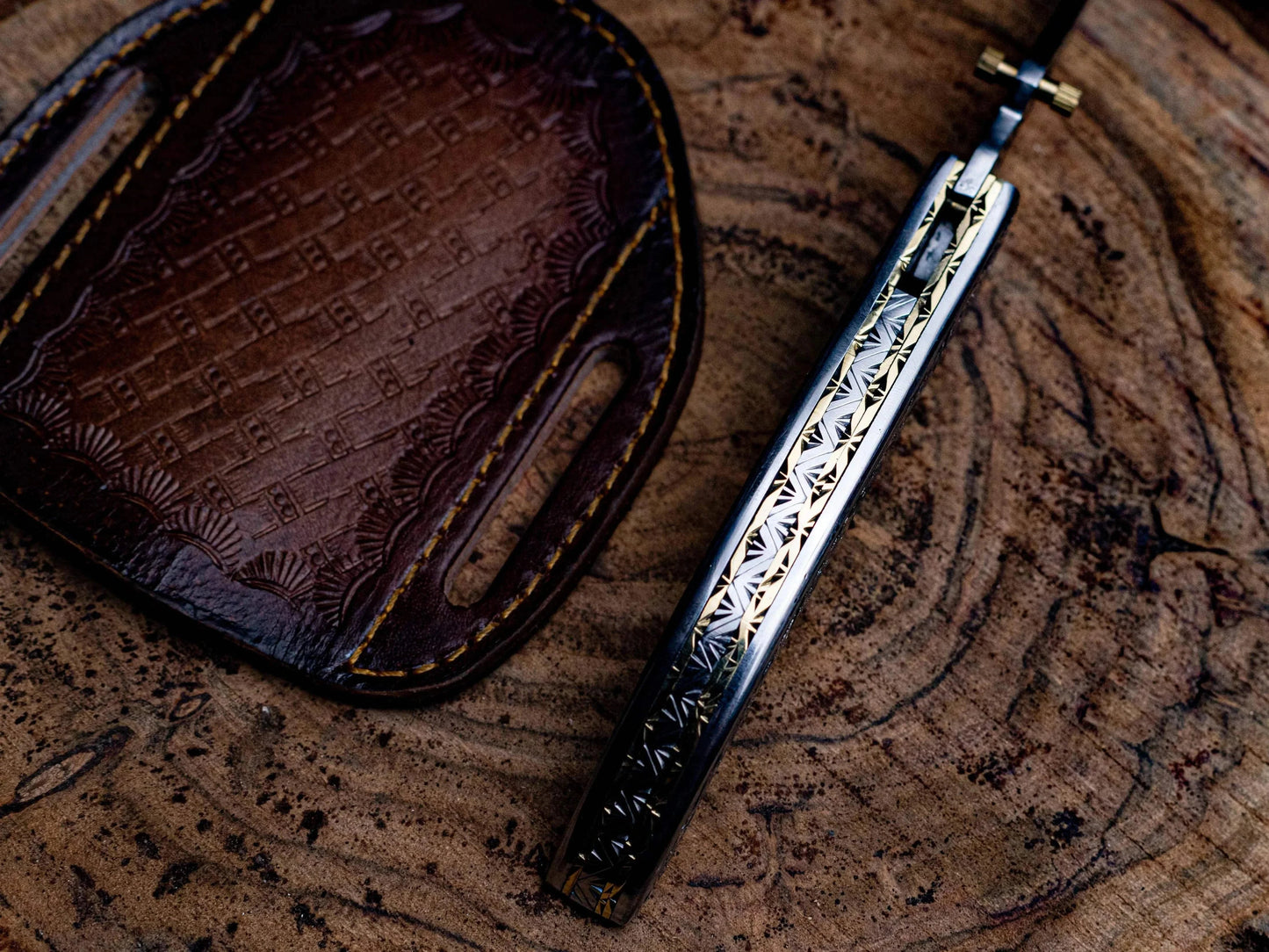 Engraved stainless steel folding knife with leather sheath on wood surface.