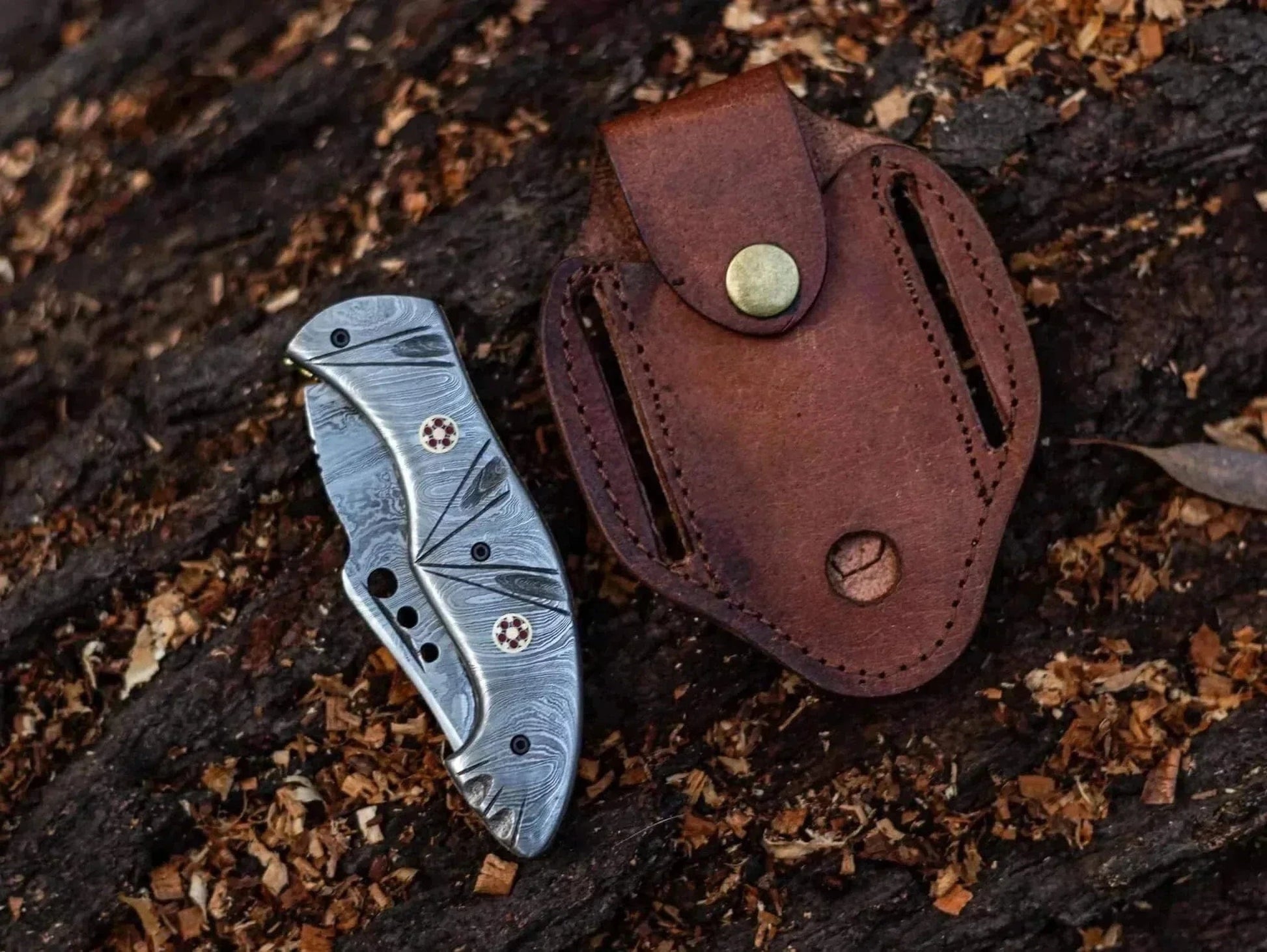Damascus Steel Folding Knife with leather sheath on wooden surface.