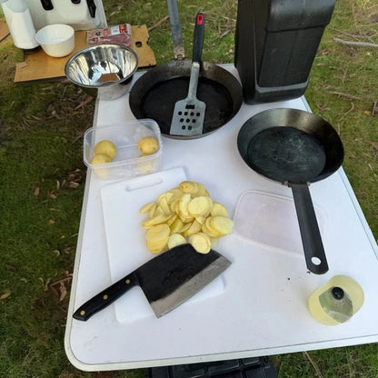 Camping cooking set with cleaver knife, sliced potatoes, and pans on outdoor table.