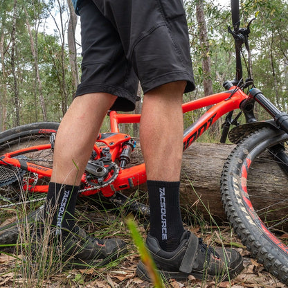 TacSource Active Train 2.0 Socks on a cyclist next to a red mountain bike in a forest setting.