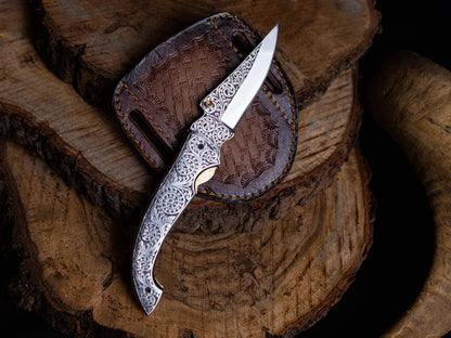 Engraved stainless steel folding knife with leather sheath on wood surface.