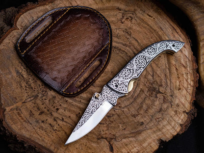 Engraved premium utility folding knife with stainless steel blade and leather sheath on a wooden background.