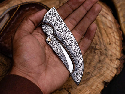 Engraved stainless steel folding knife with leather sheath lying on a wooden background.