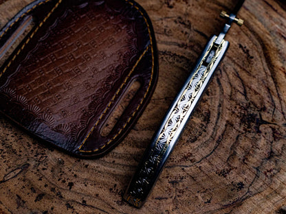 Engraved stainless steel folding knife with leather sheath on wood surface.