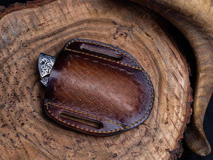 Stainless steel folding knife with engraved handle in leather sheath on wooden surface.