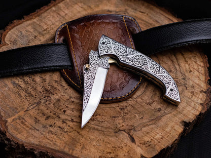 Engraved stainless steel premium utility folding knife with leather sheath on wooden background.