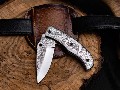 Engraved stainless steel folding knife with leather sheath on wooden background.