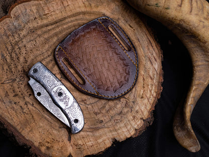 Stainless steel folding knife with engraved handle and leather sheath on wooden surface.