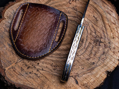 Engraved stainless steel folding knife with leather sheath on wooden surface.