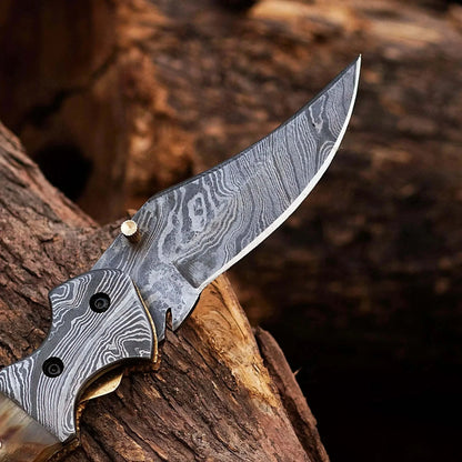 Damascus Steel Folding Knife with intricate blade design and Ram White Horn handle, displayed on wood background.