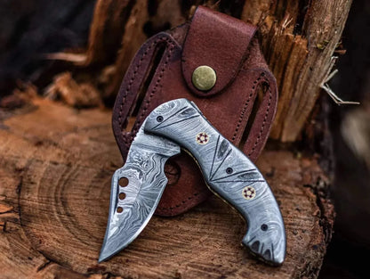 Damascus steel folding knife with intricate patterns, leather sheath, outdoor setting.